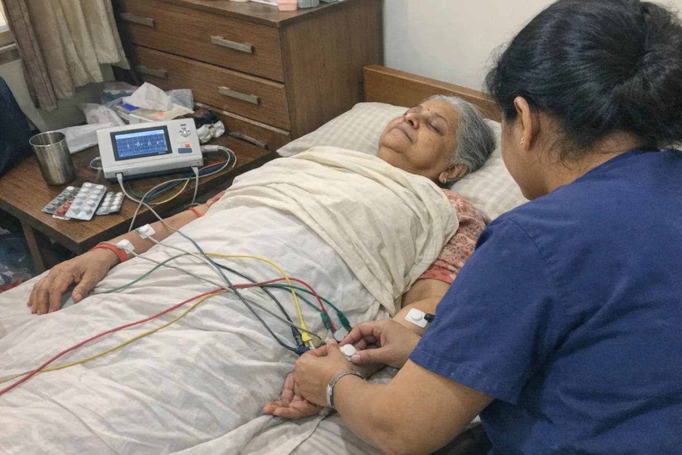 A technician attaching ECG leads to an older woman lying on her bed, with a portable ECG machine on the bedside table